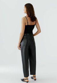 Frau mit schulterlangem, lockigem Haar, die ein schwarzes ärmelloses Oberteil, hochtaillierte schwarze Lederhosen und schwarze Slingback-Schuhe trägt, seitlich stehend.