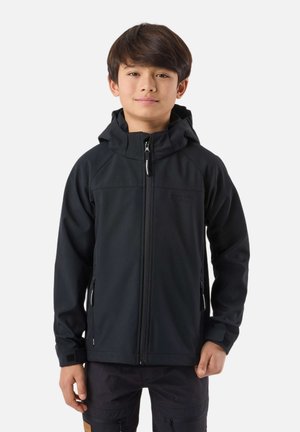 Chlapec oblečený v čiernej kapucňovej bunde na zips a tmavých nohaviciach, stojaci s uvoľnenými rukami na jednoduchom bielom pozadí.