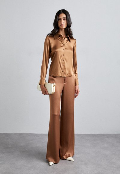 Chemise en satin marron à boutons avec col, accompagnée d'un pantalon large assorti et d'une pochette blanche. Des talons aigus crème complètent l'ensemble.