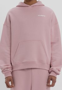 Personne portant un sweat à capuche et un pantalon de survêtement rose poussiéreux assortis avec le logo « lowlights » sur la poitrine et le pantalon, debout devant un fond uni.