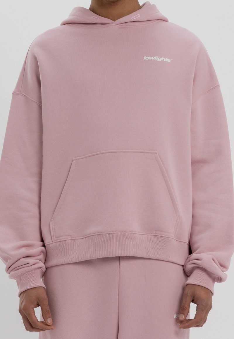 Personne portant un sweat à capuche et un pantalon de survêtement rose poussiéreux assortis avec le logo « lowlights » sur la poitrine et le pantalon, debout devant un fond uni.