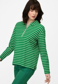 Polo a maniche lunghe verde e bianco a righe con colletto zip e spacchi laterali, abbinato a pantaloni verdi.