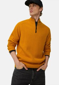 Oranger gestreifter Pullover mit halbem Reißverschluss am Kragen und schwarzen Akzenten an den Bündchen. Kombiniert mit dunkelgrauen Jeans im lässigen Schnitt.