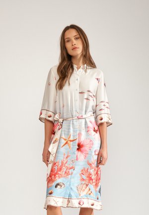 Nő, aki öves ingruhát visel háromnegyedes ujjakkal, tengeri témájú mintával, amely korallokat, kagylókat, tengeri csillagokat, halakat és nagy rózsaszín virágokat ábrázol.