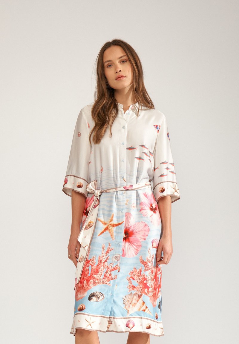 Femme portant une robe-chemise ceinturée avec des manches 3/4, présentant un motif marin composé de coraux, coquillages, étoiles de mer, poissons et grandes fleurs roses.