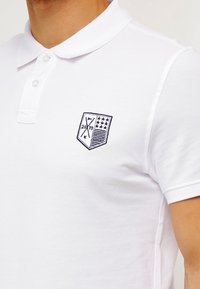 Camisa polo blanca hecha de tejido texturizado, con cuello, tapeta de botones y un logotipo bordado en forma de escudo con diseño geométrico y estrellas.