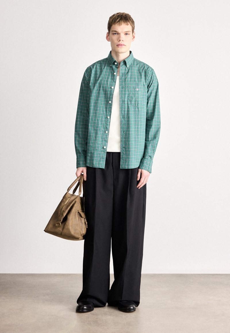 Green plaid button-up shirt, black wide-leg trousers, and brown leather bag. Model stands with neutral expression on a textured light background.