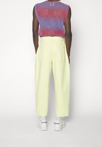 Hombre con pantalones de tiro alto en color amarillo pálido, zapatillas blancas con el texto "ICEBERG" y una parte superior sin mangas de punto rojo y morado, de espaldas.