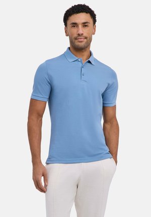 Uomo che indossa una polo a maniche corte azzurro chiaro e pantaloni bianchi, in piedi con una mano in tasca davanti a uno sfondo bianco.