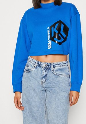 Femme portant un sweat-shirt court bleu avec un logo noir et un texte blanc, associé à un jean taille haute bleu clair.