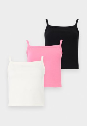 Γυναίκα με ροζ τοπ και σορτς στέκεται στο ένα πόδι φορώντας μαύρες μπαλαρίνες μπροστά σε απλό λευκό φόντο.