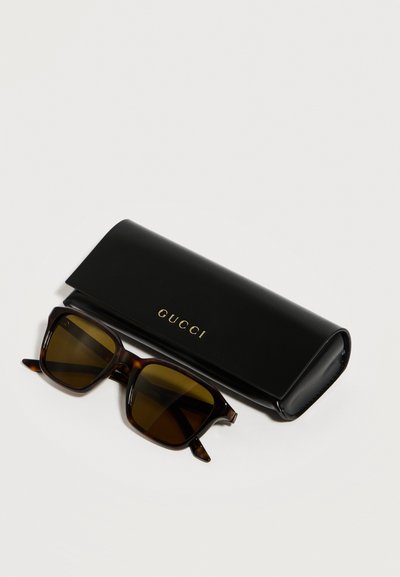 Lunettes de soleil rectangulaires en écaille de tortue avec des verres ambrés, posées à côté d'un étui noir Gucci avec des lettres dorées sur une surface blanche.