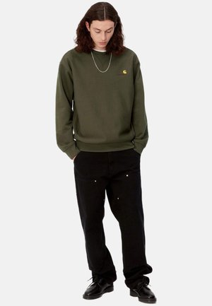 Sudadera de color verde oliva con puños y dobladillo acanalados, con un pequeño logo. Combinada con pantalones negros y zapatos negros, mostrando un ajuste relajado.