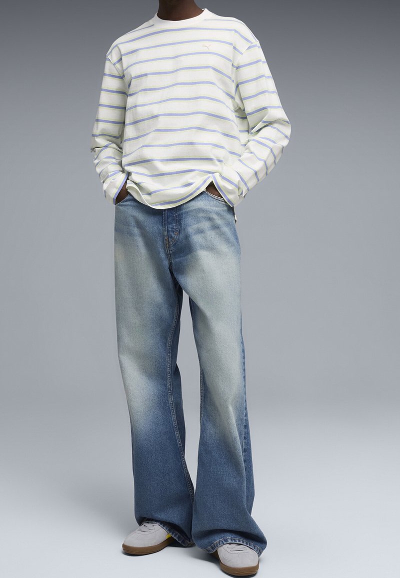 Personne portant un jean bleu délavé à jambes larges, des baskets blanches et une chemise blanche à manches longues avec de fines rayures horizontales bleues et jaunes.