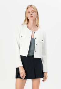 Weiße, strukturierte, kurz geschnittene Jacke mit rundem Ausschnitt, vier Fronttaschen und silbernen Knöpfen. Getragen über einem grauen Oberteil mit einem plissierten schwarzen Rock.