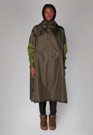 Vrouw draagt een lange olijfgroene regenponcho met capuchon en groene mouwen, zwarte broek en bruine veterwinterlaarzen, staande tegen een effen achtergrond.