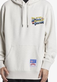 Light beige hoodie in cotton blend, featuring a graphic logo on the left chest, a front pocket, and a blue and red stitched label on the hem.