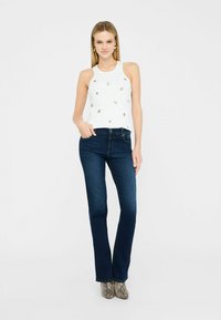 Vaqueros oscuros de campana con cintura ajustada, combinados con una blusa sin mangas blanca decorada con pequeños acentos dorados.