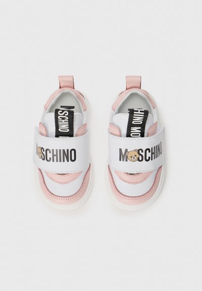 Baskets pour enfants en matériau synthétique blanc et rose clair, dotées d'une large sangle ajustable avec un texte noir et un logo d'ours.