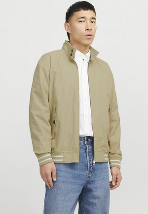 Lys beige jakke med høj krave, lynlås foran og to sidelommer. Har gråstribede manchetter og en glat, tekstureret overflade.
