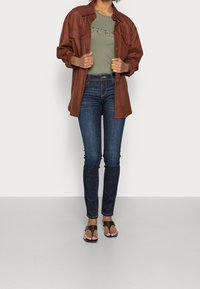 Camisa chaqueta oversize marrón, camiseta ajustada verde oliva con el texto "QUEST", jeans ajustados azul oscuro y chanclas marrones.