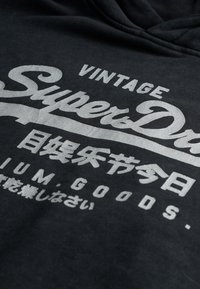 Sudadera negra con gran impresión plateada y texto. El material parece suave con una superficie ligeramente texturizada. Diseño clásico con cordones.
