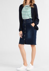 Džínová sukně v tmavě modré barvě se zapínáním na knoflíky vpředu, dvě boční kapsy, nošena se zeleným pruhovaným tričkem a bílými teniskami.