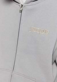 Szara bluza z kapturem na zamek, wykonana z miękkiego materiału, z zamkiem z przodu, prążkowanymi mankietami i dekoracyjnym haftowanym logo "MULTIPLY" w kolorze kremowym.