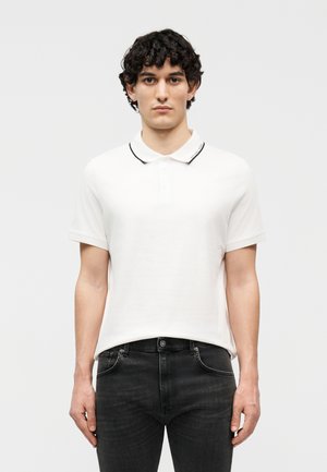 Junger Mann mit lockigem dunklem Haar, der ein weißes kurzärmeliges Poloshirt und schwarze Jeans trägt und vor einem schlichten hellen Hintergrund steht.