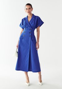 Mėlyna linine suknelė su apykakle, trumpomis rankovėmis, dvigubos sagos uždarymu, šonine skela ir kišenėmis. Dėvima su baltomis kulkšnies bateliais.