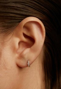 Pendiente de aro dorado con tres pequeñas gemas transparentes. Visible en el lóbulo de la oreja con el cabello castaño cubriendo parcialmente la oreja. Textura suave.