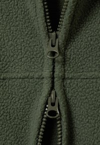 Material polar verde cu fermoar dublu cu glisieră parțial deschisă, evidențiind textura materialului și capsele de plastic ale fermoarului în prim-plan.