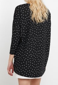 Schwarzes Shirt mit Dreiviertelärmeln, das ein verstreutes Muster aus kleinen Kreisen in Weiß und Blau zeigt. Weicher Stoff mit lockerer Passform.