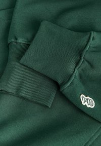Poignets de sweat-shirt vert foncé avec texture côtelée et petit logo blanc brodé sur la manche près du poignet.