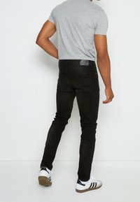 Jeans negros ajustados con bolsillos traseros, costuras sutiles y parche de marca de cuero. Combinados con una camiseta de manga corta gris claro y zapatillas.