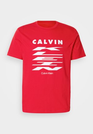 Rdeča bombažna majica s kratkimi rokavi, ki ima bold belo besedilo "CALVIN KLEIN" in diagonalni črtasti vzorček. Okrogla ovratnica.