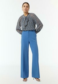 Blauwe bloemenblouse met een gerimpelde halslijn, gecombineerd met hoge taille, wijde blauwe broek. De outfit is afgewerkt met metallic haksandalen.