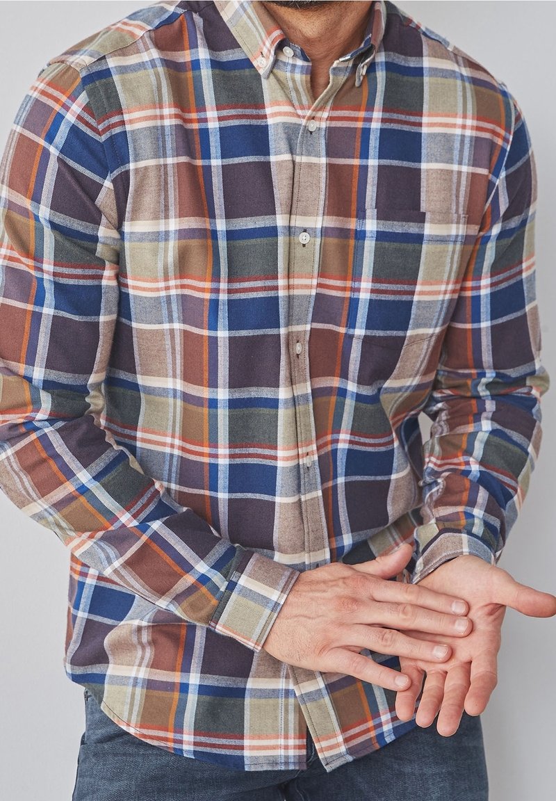 Homme portant une chemise à boutons à carreaux multicolores se frotte la paume gauche avec la main droite sur un fond uni.