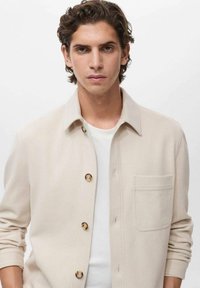 Veste chemise beige clair à col fabriquée en tissu doux ; elle dispose de boutons dorés, d'une poche plaquée et de manches côtelées.