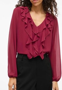Bordowa bluzka z lekkiego, przezroczystego materiału, z dekoltem w serek i falbankami na przodzie oraz rękawach. W zestawie z czarnymi spodniami.