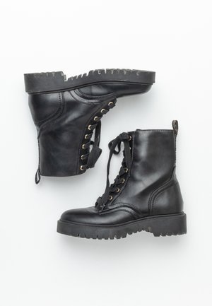 Bottes de combat en cuir noir avec semelles crantées épaisses et lacets noirs, une botte debout et l'autre sur le côté sur fond blanc.