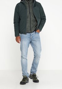 Chaqueta de plumas verde oscuro con capucha, superpuesta a un suéter gris con cremallera. Unos jeans azul claro y unos zapatos de senderismo verdes completan el conjunto.