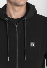 Sudadera con capucha negra, con cremallera y textura acanalada, que presenta un parche de marca y detalles de cremallera metálica. Los cordones ajustables completan el diseño.