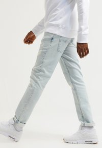 Person walking wearing light blue ripped jeans, white sneakers, white socks, and a white long-sleeve shirt against a plain background.