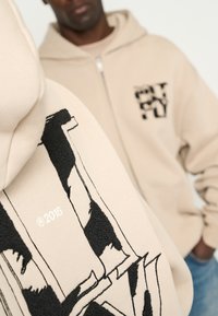 La sudadera con cremallera beige presenta bordados y un logo en negro, con un detalle texturizado. El fondo muestra a un segundo modelo con una sudadera similar.