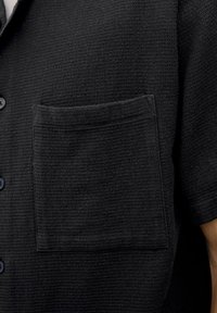 Chemise noire à manches courtes texturée avec une poche poitrine et une fermeture à boutons sur le devant, fabriquée à partir d'un tissu tissé avec un motif subtil.