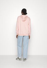 Žena s copánky ve spodním drdolu, oblečená v světle růžové mikině s kapucí, světle modrých širokých džínách a zeleno-bílých teniskách Nike, stojící zády k nám.