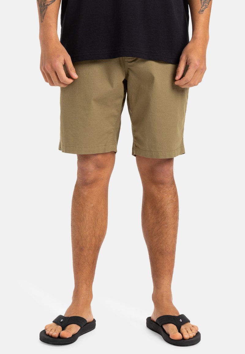 Khaki szorty o luźnym kroju, wykonane z lekkiego materiału. Noszone z czarnymi klapkami. Widoczne szwy i prosty design.