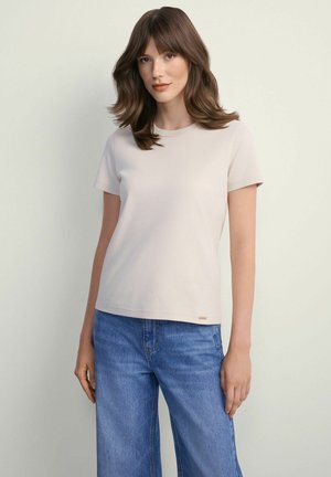 Młoda kobieta z brązowymi włosami do ramion, ubrana w jasnobeżowy t-shirt z krótkim rękawem i niebieskie jeansy z wysokim stanem na jednolitym tle.