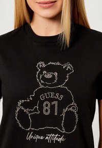 Czarny t-shirt z grafiką niedźwiedzia wykonaną z cyrkonii. Zawiera napis „GUESS 81” oraz „Unique attitude” w stylu kursywy pod niedźwiedziem.
